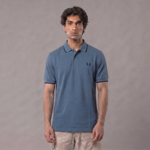A man wearing a blue short-sleeve polo shirt with a small embroidered logo on the chest, paired with beige pants, standing against a plain gray studio background | AH Urban Wear PK