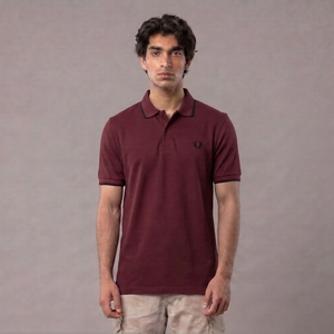A man wearing a short-sleeve polo shirt in maroon with a small embroidered logo on the chest, paired with light beige pants, standing against a plain gray studio background | AH Urban Wear PK