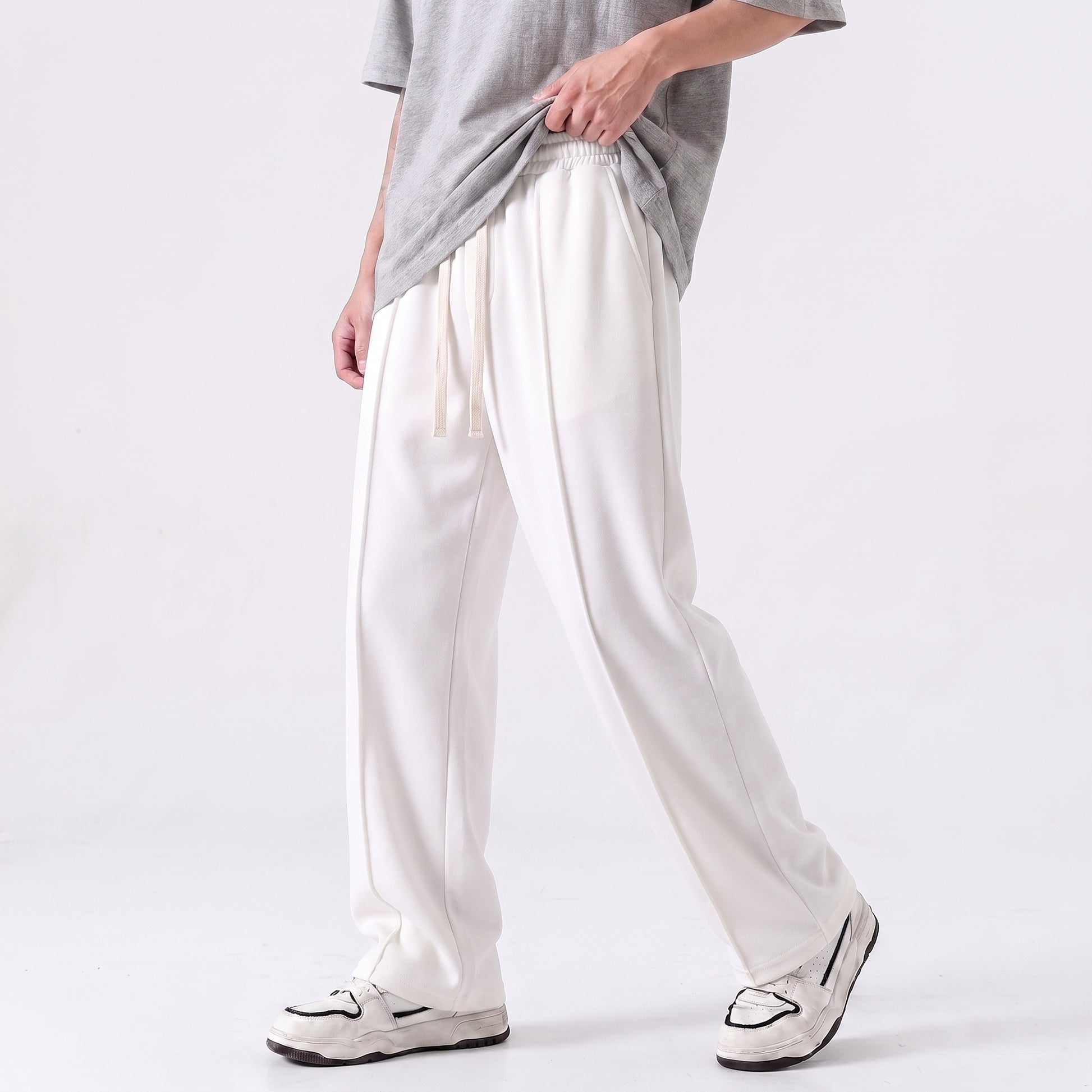 A person wearing loose white drawstring baggy trouser with a gray T-shirt and white sneakers, shown from the chest down against a clean light gray studio background l AH Urban Wear Pk